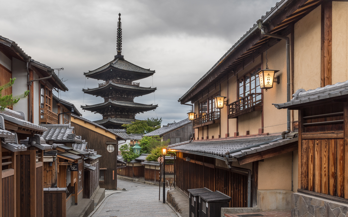 Yasaka-dori in Kyoto at first light: a stone-paved lane between wooden machiya houses, lit paper lanterns, and the five-storey Yasaka pagoda rising at the end of the street.